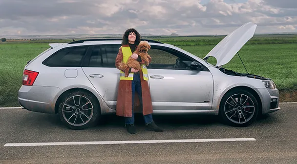 Eine Frau steht vor einem Auto mit geöffneter Motorhaube, wobei sie in die Kamera schaut und einen Hund in der Hand hält.